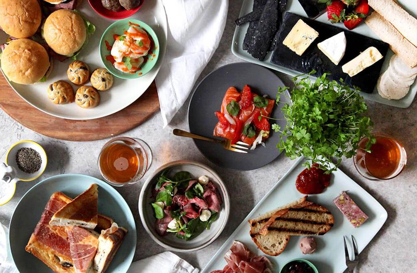 Colorful spread with burgers, salmon, cheese, breads, greens, drinks, herbs, and cookies.