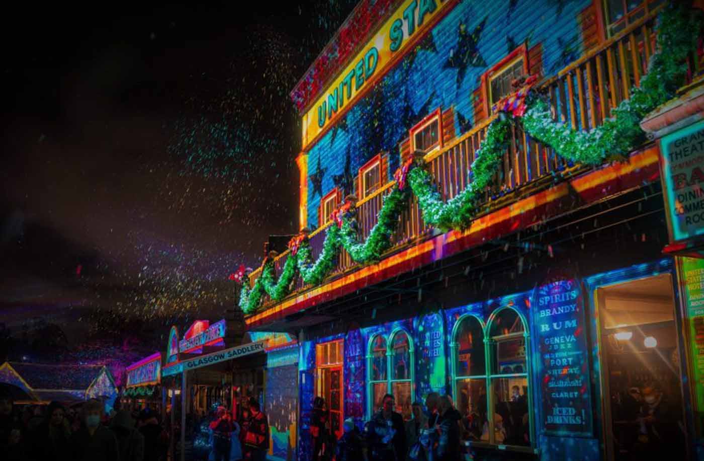 Festive street with colorful light projections, green garlands, and people gathering below.