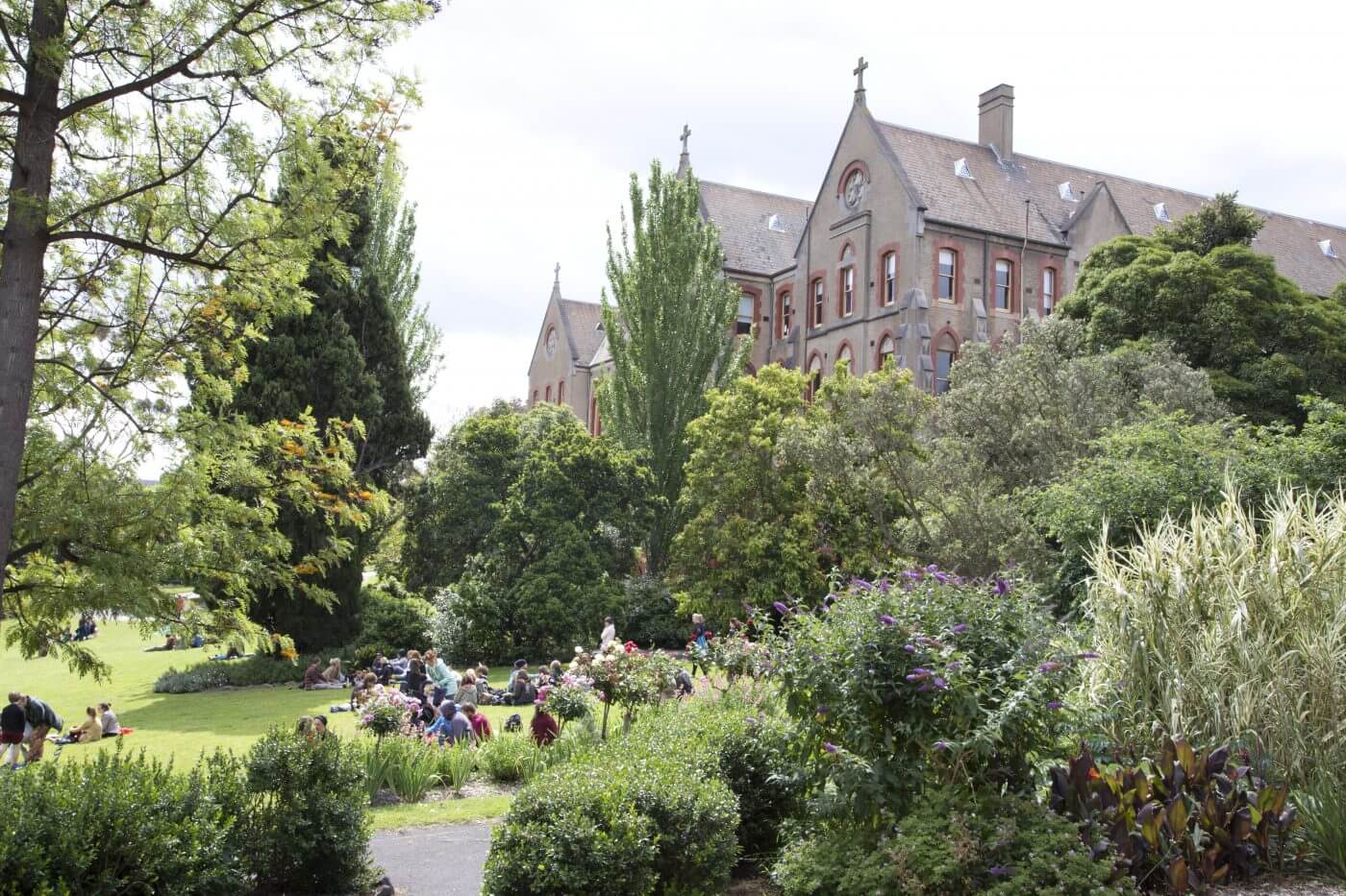 Blog Historic building with pointed roofs and gardens, as people relax on the sunny lawn nearby.