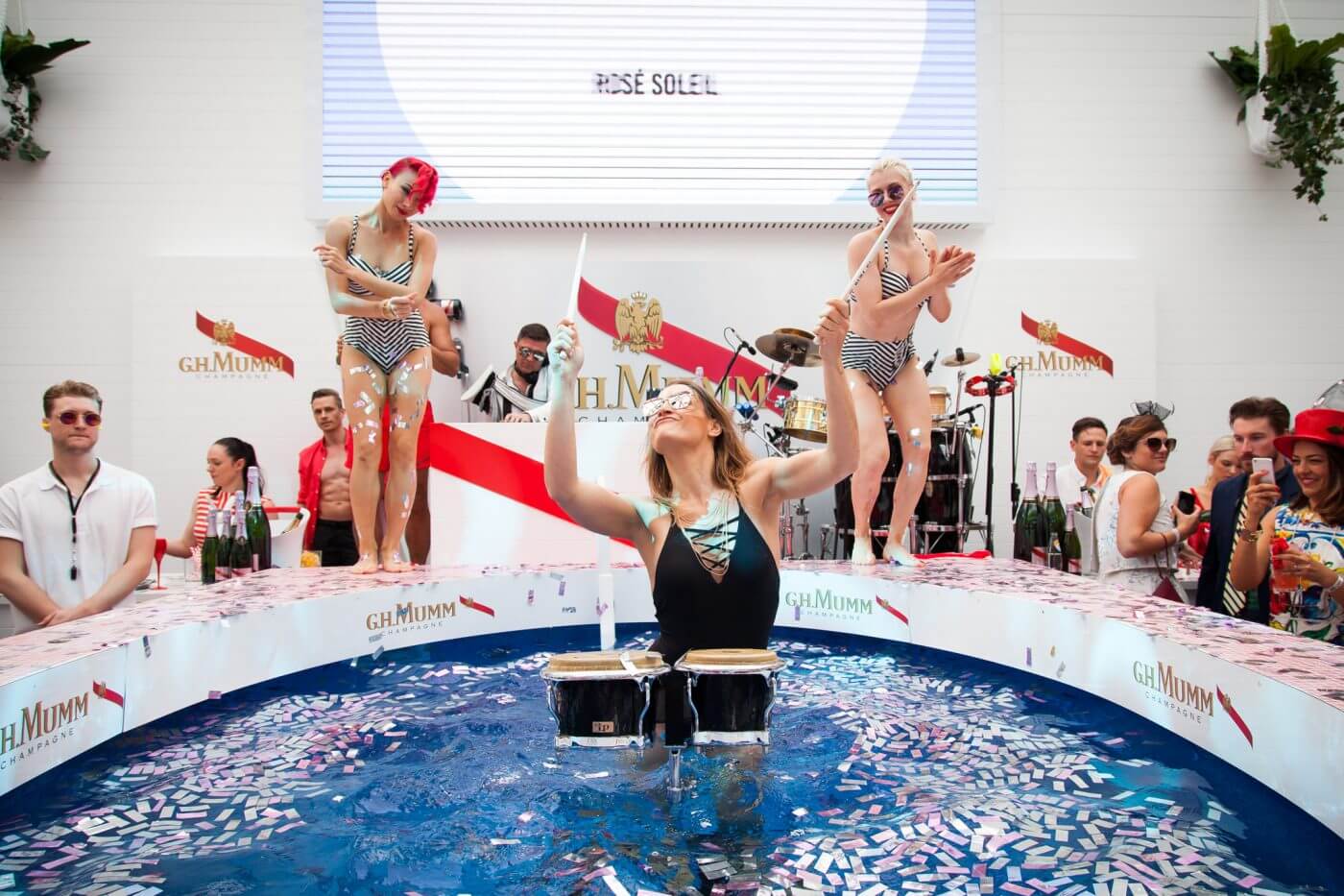 Blog Woman plays bongos in pool with confetti; dancers in stripes perform as crowd watches.