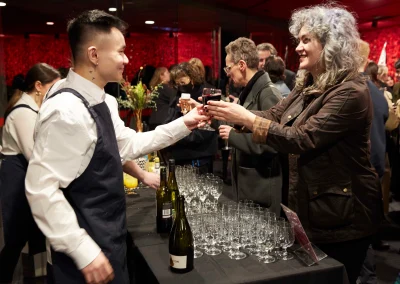 Bartender serves a drink at a lively event with guests and a table of glasses and bottles.