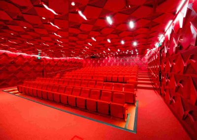 Modern auditorium with red walls and seats lit by bright overhead lights.