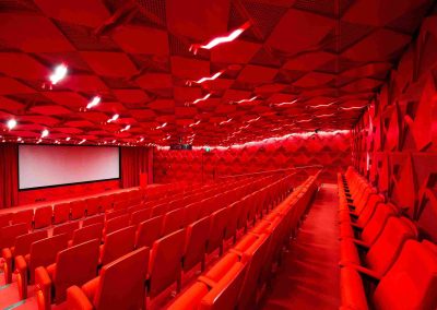 Interior of the Shrine of Remembrance auditorium with rows of red seats, red geometric walls and ceiling, facing a large projection screen at the front.