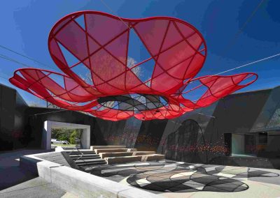 South‑east Education Courtyard at Melbourne’s Shrine of Remembrance, featuring a large red, poppy‑shaped suspended canopy casting patterned shadows over tiered seating and concrete surfaces.