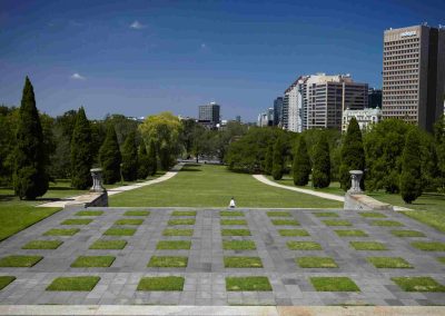 Landscaped park with lawns and trees, set against modern city buildings and clear blue sky.